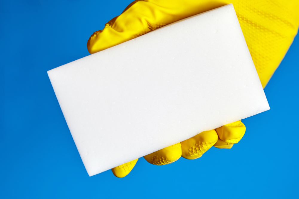 Human hand in yellow rubber glove holds a white melamine sponge on blue background