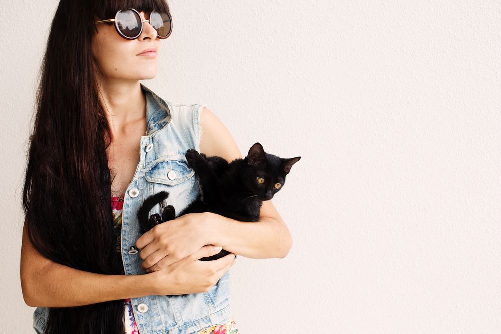Long black hair fashion girl in sunglasses holding little black cat on white wall background