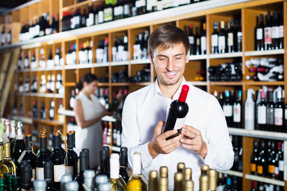 Happy young man choosing bottle of wine in shop with alcohol beverages